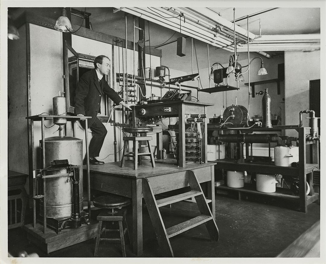 Early 20th-century photo of a man in a suit stepping out through a small door in the wall of a large respiration calorimeter, surrounded by laboratory equipment