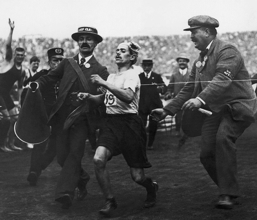 Black-and-white photo of an exhausted marathon runner wearing number 19, flanked by officials as he staggers toward the finish in a packed stadium