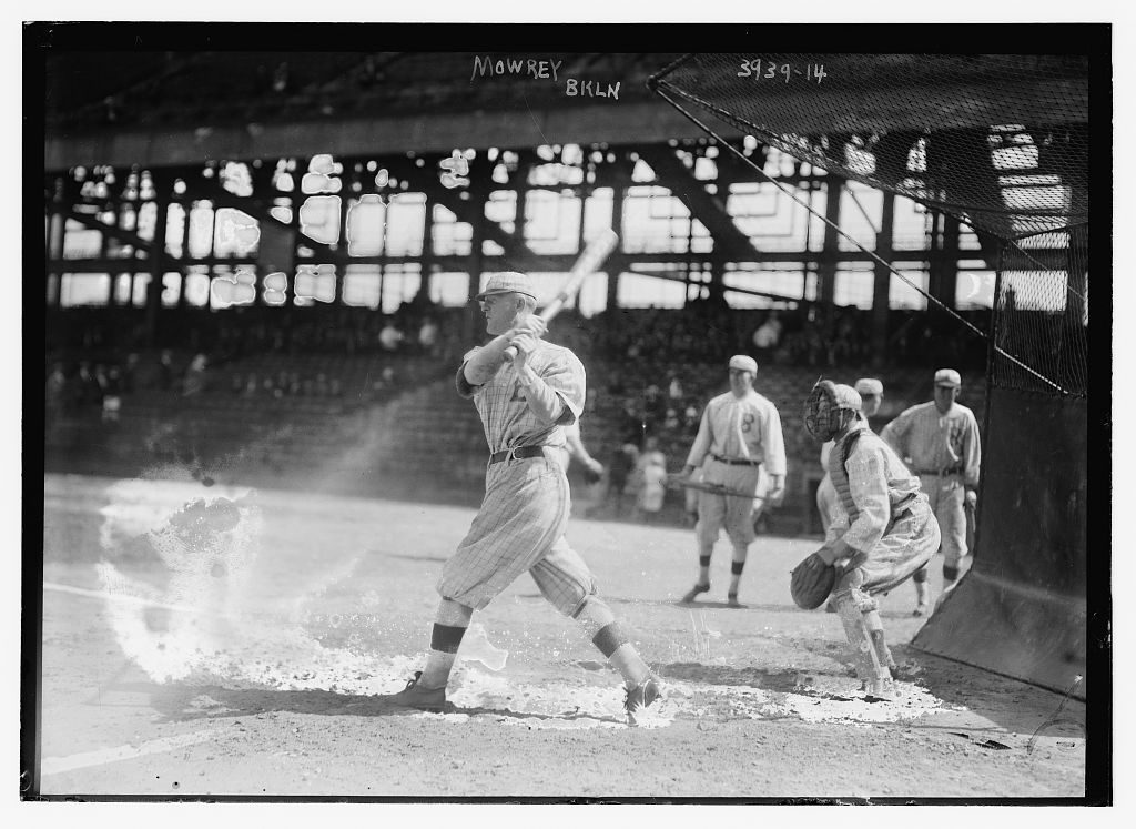 Black-and-white 1916 photograph of a baseball batter swinging, with the catcher crouched behind him